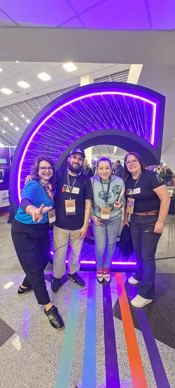 Quatro participantes do ConectaPR posam diante de uma grande estrutura circular iluminada em roxo, com faixas coloridas no piso do espaço do evento.